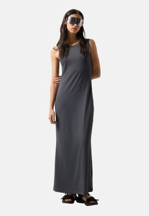 Kobieta w długiej, szarej sukience bez rękawów, dużych futurystycznych okularach przeciwsłonecznych i czarnych sandałach stoi na tle białego tła.
