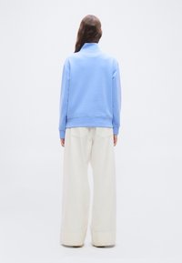 Γυναίκα που στέκεται με την πλάτη, φορώντας ανοιχτό μπλε φούτερ με ζιβάγκο και άσπρο παντελόνι φαρδιάς γραμμής σε μονόχρωμο φόντο.