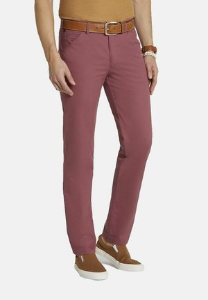 Mann trägt rosafarbene Slim-Fit-Hose mit Seitentaschen, einen braunen Ledergürtel, braune Slipper und ein braunes Oberteil mit Armbändern.