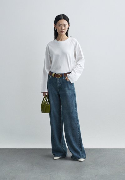 Biała koszula z długim rękawem, niebieskie szerokie dżinsy, brązowy pasek, zielona torebka oraz białe buty z ostrym czubkiem. Gładkie tekstury, codzienny styl.