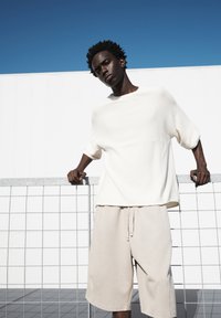 Top lavorato a maglia bianco con maniche ampie, abbinato a pantaloni beige, in piedi contro una recinzione a griglia con un cielo blu limpido sullo sfondo.