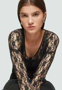 Schwarzes Spitzenshirt mit langen Ärmeln und Blumenmuster, mit rundem Ausschnitt und transparenter Textur, kombiniert mit schwarzen Hosen. Silberne Creolen.
