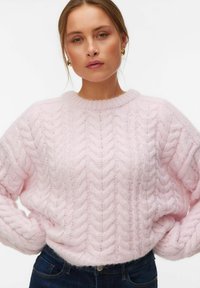 Femme portant des boucles d'oreilles créoles dorées et un pull rose à torsades rentré dans un jean bleu foncé sur un fond blanc.