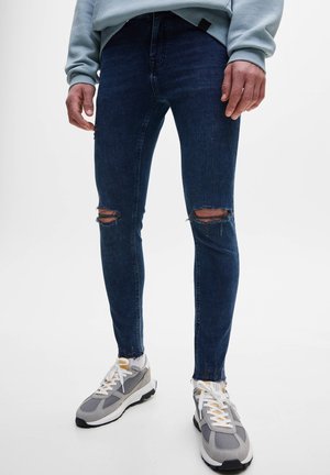 Personne portant un jean bleu foncé moulant et déchiré ainsi que des baskets grises, debout devant un fond uni.