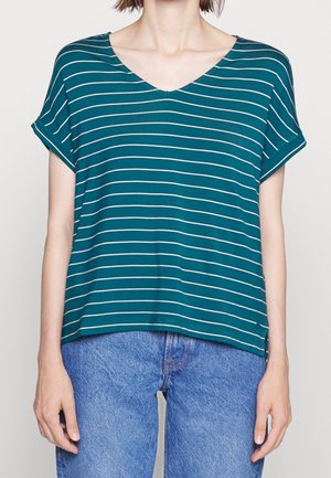 Nainen, jolla on päällään teal-värinen V-kaula-aukollinen, lyhythihainen pusero, jossa on valkoisia vaakaraitoja, ja siniset farkut, seisomassa vaaleaa taustaa vasten.