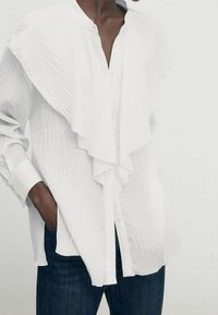 Chemise blanche boutonnée en tissu texturé, avec un col à volants drapé, des manches longues et une coupe décontractée. Associée à un denim foncé.