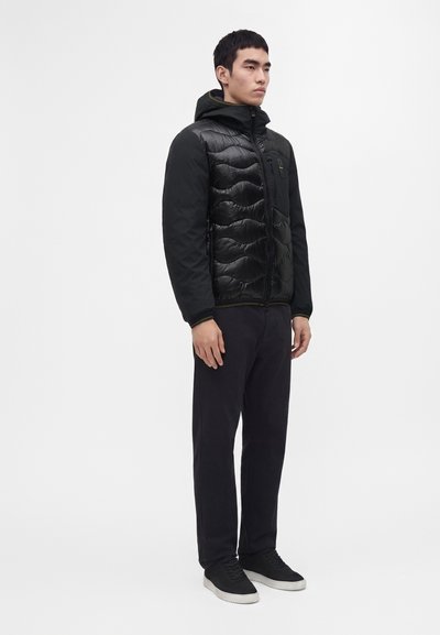 Homme debout portant une veste matelassée noire avec capuche, un pantalon noir et des baskets noires à semelles blanches sur fond blanc.
