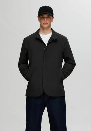 Giacca leggera nera con design a bottoni, tasche laterali e colletto, indossata sopra una camicia bianca e jeans scuri. Inclusa una berretto nero.