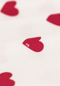 Motivi a cuore rossi su un morbido tessuto bianco, con un piccolo marchio "PB" nell'angolo inferiore. La texture del materiale appare liscia.