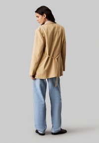 Beżowa marynarka o dopasowanym kroju z krawatem z tyłu, w połączeniu z jasnoniebieskimi spodniami typu wide-leg. Materiał wydaje się gładki, z subtelnymi detalami przeszycia.