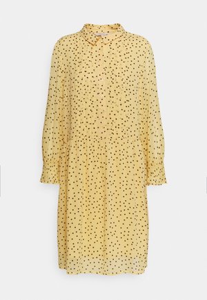 Keltainen mekko mustilla pilkuilla, kauluksellinen pääntie, nappikiinnitys edessä, pitkät hihat ja kootut vyötärö, joka tarjoaa rentoa istuvuutta.