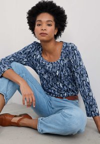 Granatowa bluzka z długim rękawem w biały kwiatowy wzór, połączona z jasnoniebieskimi dżinsami i brązowymi butami o teksturowanej powierzchni. Siedząca pozja na szarym podłożu.