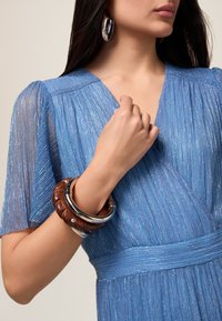 Rochie albastră cu textură plisată și mâneci scurte, având un decolteu în formă de V. Accesoriile includ o brățară din lemn legată și un bangle din argint.