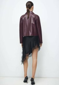 Bordowa skórzana krótka kurtka zestawiona z czarną asymetryczną sukienką z koronkową lamówką oraz czarnymi szpilkami.