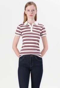 Νεαρή γυναίκα με ίσια μαλλιά, φορώντας λευκό και μπορντώ ριγέ πόλο και σκούρους μπλε τζιν, στέκεται με τα χέρια πίσω από την πλάτη.