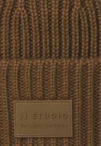 Bruin gebreid stof met een rechthoekig gestikt leren label met de inscriptie "JJ STUDIO Our Legacy Your Future."