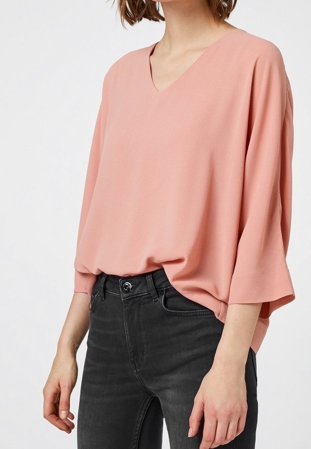 Blusa de color rosa claro, con cuello en V, hecha de una tela suave y con mangas anchas, llevada sobre jeans de color gris oscuro. La parte superior tiene un corte holgado y una textura suave.