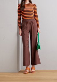 Gestreepte langemouwen top in oranje en bruin, bruine wijde broek, oranje blokhak sandalen en een groene handtas.