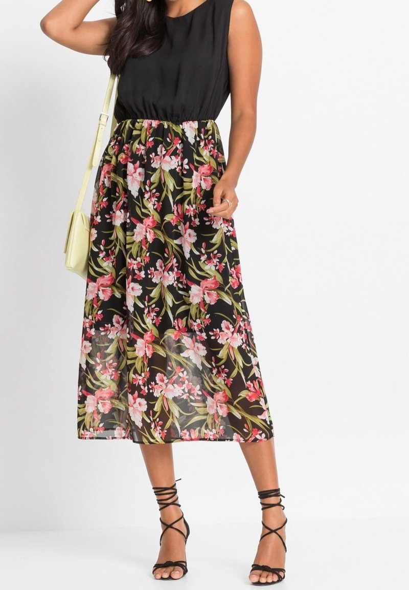 Černý top bez rukávů s průsvitnou černou sukní s květinovým potiskem v růžové a zelené barvě; modelka má na nohou černé sandály s pásky.