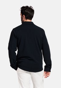 Chemise noire à manches longues avec un col et une coupe droite. Présente des détails de couture et un tissu lisse et texturé.