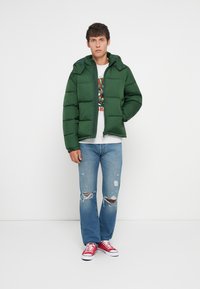 Veste matelassée verte avec une fermeture éclair et une capuche, portée sur un t-shirt graphique gris. Associée à un jean bleu déchiré et des baskets rouges.