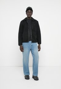 Black fleece jacket with a zip front and pocket, worn over a black t-shirt, paired with light blue jeans and dark sneakers.