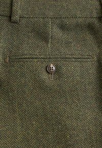Grön herringbone-mönstrad tyg med texturerad yta. Har en enkel knäppning vid bakfickan som ger en detalj till designen.