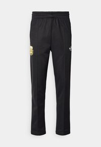 Pantalons de survêtement noirs avec une taille élastique, présentant un détail de blason jaune sur la cuisse gauche et un logo Adidas blanc sur le côté droit.