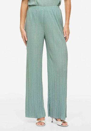 Nő magasderekú, bőszárú zsályazöld pliszírozott nadrágban, fehér, nyitott orrú, kis sarokkal rendelkező szandált viselve, fehér háttér előtt állva.