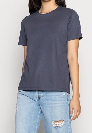Femme portant un t-shirt gris foncé uni à manches courtes et un jean bleu clair usé, debout devant un fond uni.