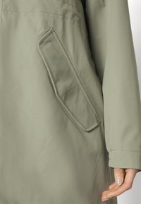 Veste vert kaki avec une poche latérale à rabat, tissu lisse et coupe ajustée. Coutures visibles le long des coutures.