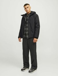 Chaqueta negra impermeable con capucha, bolsillos con cremallera, llevada sobre una camisa a cuadros. Combinada con pantalones cargo negros y zapatillas con detalles en gris.