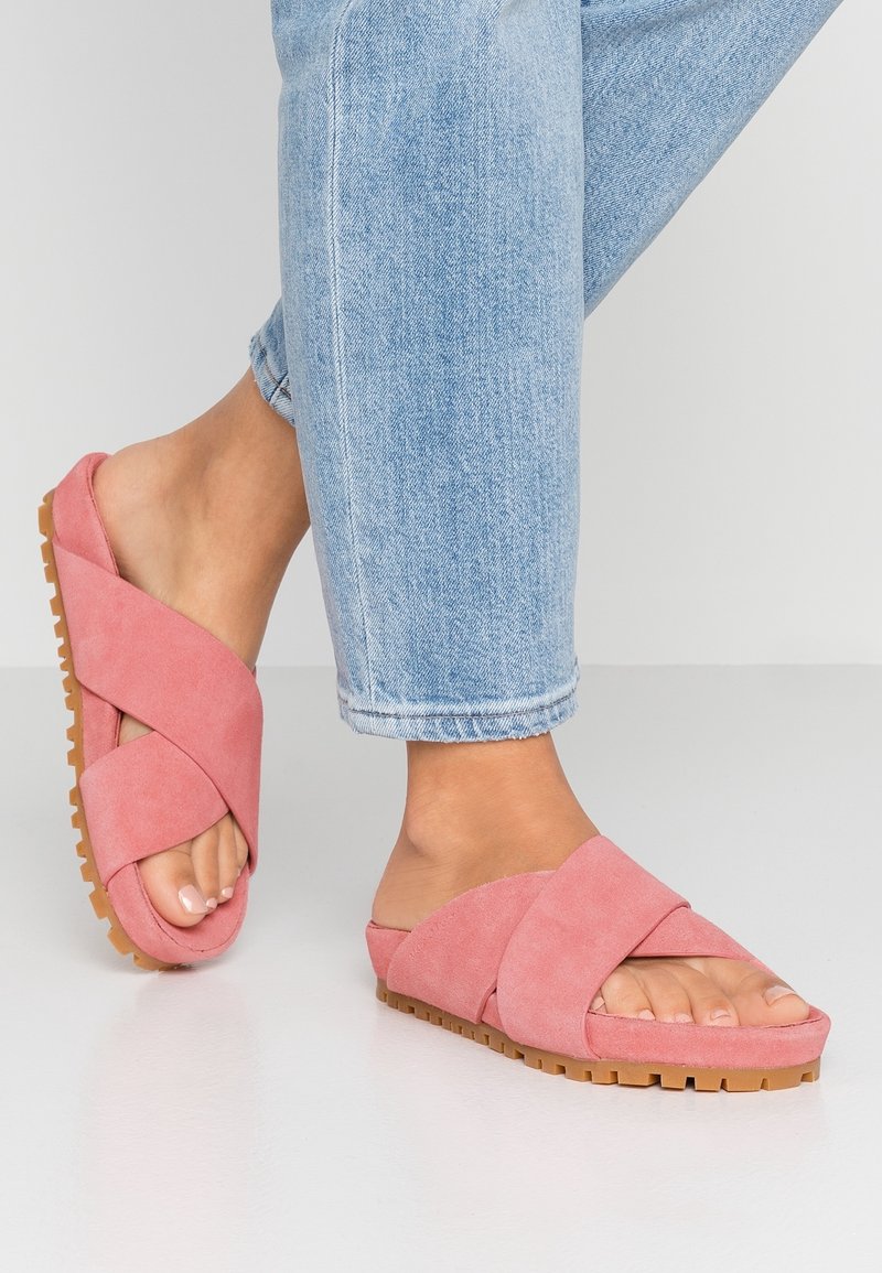 Sandales à glissière en daim rose avec un design de sangle croisée et une semelle en caoutchouc texturé beige, portées avec du denim bleu clair.