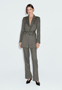 Donna in piedi che indossa un blazer grigio con cintura e pantaloni a palazzo abbinati, con tacchi a punta neri, su uno sfondo bianco uniforme.