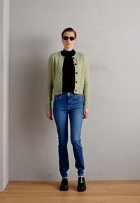 Groene gebreide cardigan over een zwarte coltrui, blauwe spijkerbroek en zwarte instappers; model draagt donkere zonnebril. Eenvoudig, getailleerd ontwerp.