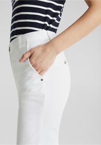 Pantalon en coton blanc à coupe slim, avec poches avant et boutons métalliques. Le mannequin porte un haut rayé bleu marine et blanc.