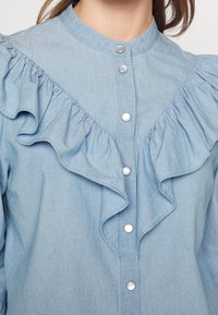 Blouse bleu clair à volants avec boutons pression et col rond haut portée par une personne aux cheveux bruns.