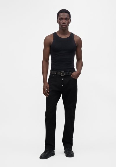 Jeune homme portant un débardeur noir, un pantalon noir avec ceinture et des chaussures noires, debout devant un fond blanc uni.