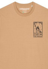 Beige t-shirt gemaakt van zachte stof, met een vierkant zwart grafisch ontwerp van de tarotkaart "De Tovenaar" op het linkerborstgebied.