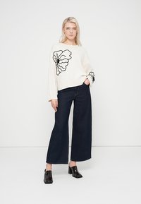 Bílý pletený svetr s černou květinovou výšivkou na levém rameni; kombinovaný s džínami s vysokým pasem, tmavě modrými s širokými nohavicemi, a černými mule botami.