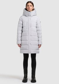 Šviesiai pilkas pūkinis paltas su aukštu apykakliu, priekiniais spaudžiamosiomis sagomis ir dėžutės formos siluetu. Yra krūtinės kišenė, dėvimas kartu su juodomis tamprėmis ir batais.