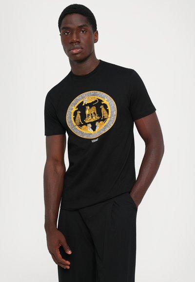Bărbat îmbrăcat cu un tricou negru, cu modele de feline mari brodate în aur și argint, și pantaloni negri, stând cu o mână în buzunar.