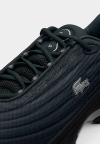 Baskets texturées noires avec détails en mesh, lacets plats et un logo gris subtil sur le côté. Présentent un design élégant et moderne ainsi qu'une forme arrondie.