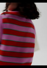 Personne aux cheveux bouclés foncés tournant le dos, portant un pull violet avec de larges rayures horizontales rouges et fines rayures vertes sur un fond uni.