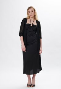 Schwarzes Kleid mit tailliertem Oberteil, drapiertem Stoff und einem runden Akzent. Kombiniert mit einer durchsichtigen, offen gestrickten schwarzen Strickjacke und schwarzen Absätzen.