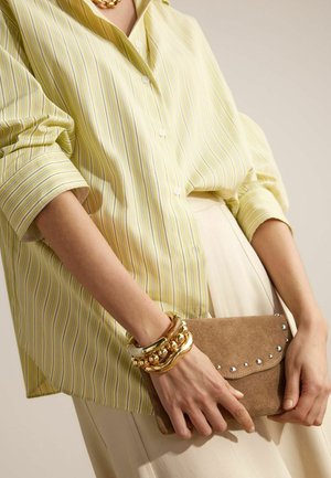 Personne portant un chemisier ample à rayures jaunes et un pantalon crème, tenant une pochette en daim marron avec des clous métalliques, et des bracelets dorés épais au poignet.