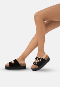 Sandales à plateforme noires avec une base en liège texturé, une tige en daim et des boucles décoratives ornées de pierres de couleur claire.