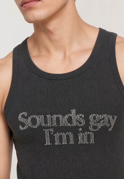 Gros plan d'une personne portant un débardeur côtelé noir avec des clous argentés formant le texte "Sounds gay I'm in" à l'avant.