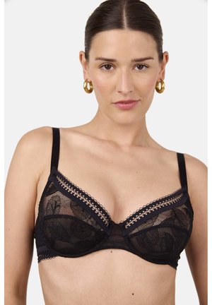 Vrouw met donker haar naar achteren gekamd, die een zwarte kanten beha en gouden oorringen draagt, kijkt recht vooruit tegen een witte achtergrond.