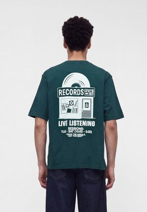 Homme portant un t-shirt vert foncé avec un graphisme blanc d'un magasin de disques et un texte sur les séances d'écoute en direct dans le dos.